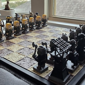 May include: A detailed chess set with themed pieces. The pieces are arranged on a checkered board with a dark border. The pieces on one side have blonde hair and black outfits, while the opposing pieces are black. The board is set on a gray surface.