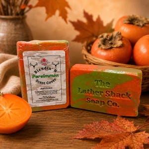 May include: Two rectangular bars of handmade soap with orange and green swirls. One bar features a white label with the text Blendin' N Persimmon Scent Cover, while the other bar has The Lather Shack Soap Co. embossed on the front surface.