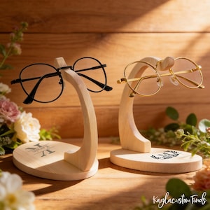 May include: Two wooden eyeglasses stands, one with black-framed glasses and the other with gold-framed glasses. The stands are light-colored wood with a curved design. One stand has a monogram, and the other has a decorative design.