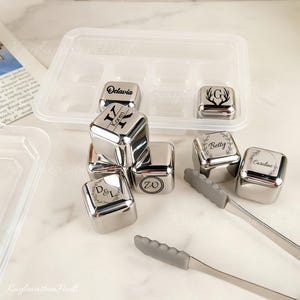 Pode incluir: Uma coleção de cubos de gelo personalizados em aço inoxidável com nomes e iniciais gravados. Os cubos são exibidos com uma bandeja de cubos de gelo de plástico e um par de pinças com cabo cinza. Os nomes "Octavia", "Karen", "Betty" e "Caroline" são visíveis.