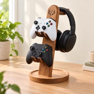 May include: A wooden gaming stand with a circular base and vertical support featuring an engraved letter C inside a deer antler design. The stand holds one white and one black game controller on front hooks and black headphones on a side hook.