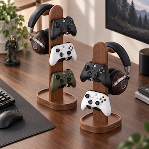 May include: Two vertical wooden gaming stands with oval bases, each holding one pair of over-ear headphones and multiple game controllers. The left stand holds three controllers in black, white, and olive green, while the right holds two in black and white.