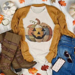 Op de afbeelding: Een herfstachtige compositie met een wit t-shirt met een patchwork pompoenontwerp, een mosterdgeel vest, een blauwe jeans, bruine kniehoge laarzen en decoratieve pompoenen en bladeren. Zonnebril en telefoon zijn ook inbegrepen.