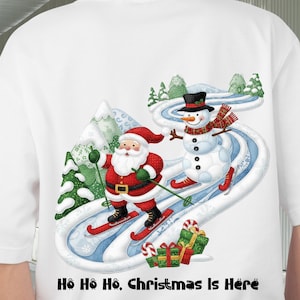 May include: White t-shirt featuring a festive Christmas design. Santa Claus and a snowman are skiing down a snowy slope. The text "Ho Ho Ho, Christmas Is Here" is printed at the bottom. A fun and colorful holiday-themed tee.