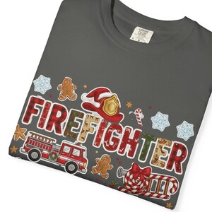 May include: A folded charcoal gray t-shirt with a festive graphic. The design includes a red fire truck, a firefighter's helmet, gingerbread men, snowflakes, and the word "FIREFIGHTER" in a plaid and patterned font. The shirt is a Comfort Colors brand.