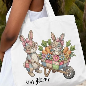 May include: White tote bag with a colourful illustration of two bunnies with glasses, carrots, and flowers in a wheelbarrow. The text "STAY HOPPY" is printed below the illustration. The bag is carried by a person on a beach.