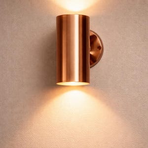 May include: A cylindrical copper wall sconce with a warm, inviting glow. The fixture is mounted on a circular base and casts light upwards and downwards, creating a cozy ambiance. Ideal for interior lighting.