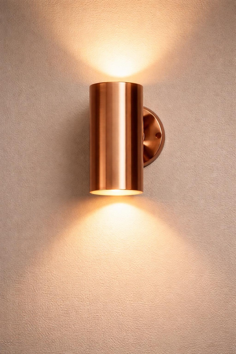 May include: A cylindrical copper wall sconce with a warm, inviting glow. The fixture is mounted on a circular base and casts light upwards and downwards, creating a cozy ambiance. Ideal for interior lighting.