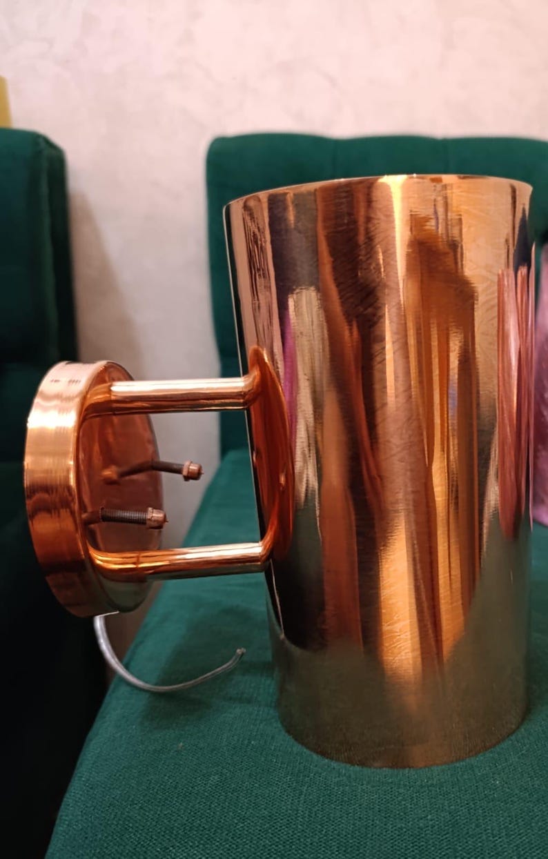 May include: A copper-colored wall sconce with a cylindrical shade and a bracket for mounting. The sconce has a polished finish, reflecting the light. The fixture is designed for indoor use and adds a touch of elegance to any room.