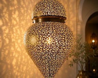 May include: A hanging pendant lamp with a teardrop-shaped metal shade featuring an intricate, laser-cut filigree pattern. The brass-toned finish glows from an internal light source, casting detailed, complex shadows on the surrounding wall.