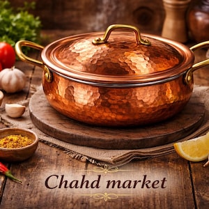 Puede incluir: Una olla de cobre con tapa y asas doradas está sobre una superficie de madera. La olla tiene una textura martillada. Alrededor de la olla hay tomates, especias, ajo y una rodaja de limón. El texto "Chahd market" es visible.