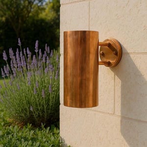 May include: A cylindrical outdoor wall sconce made of copper, mounted on a light-colored stone wall. The sconce has a round backplate and two small arms connecting to the cylindrical shade. The surrounding area includes lavender and green foliage.