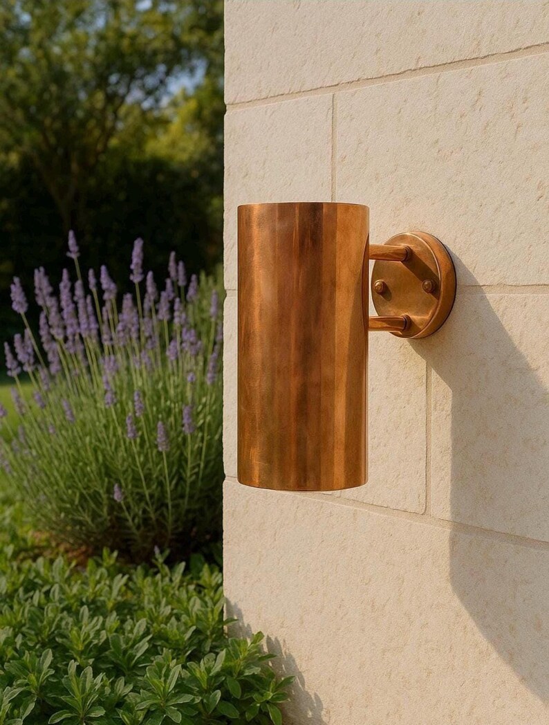 May include: A cylindrical outdoor wall sconce made of copper, mounted on a light-colored stone wall. The sconce has a round backplate and two small arms connecting to the cylindrical shade. The surrounding area includes lavender and green foliage.
