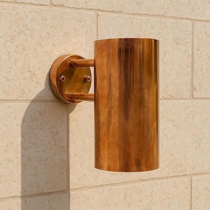 May include: A copper-colored wall sconce with a cylindrical shade and a circular mounting plate. The sconce is attached to a light-colored, textured stone wall. The fixture has a warm, metallic finish.