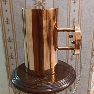 May include: A copper-colored wall sconce with a cylindrical body and a circular mounting plate. The sconce is mounted on a dark brown wooden pedestal. The pedestal has a decorative, turned column and a round top. A wire extends from the sconce.