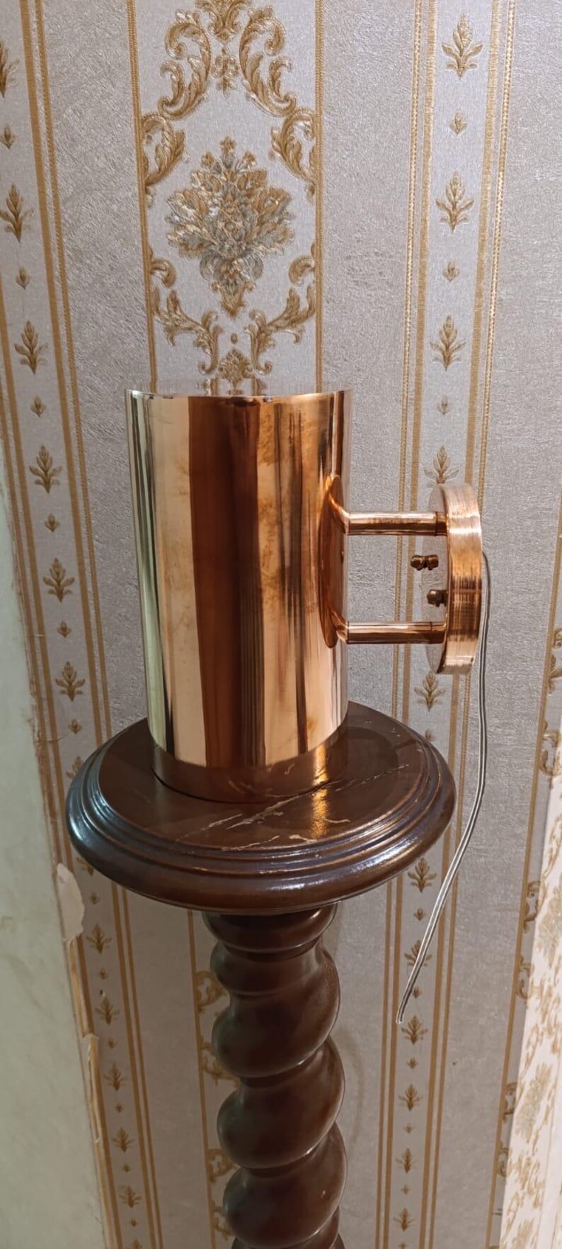 May include: A copper-colored wall sconce with a cylindrical body and a circular mounting plate. The sconce is mounted on a dark brown wooden pedestal. The pedestal has a decorative, turned column and a round top. A wire extends from the sconce.