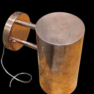 May include: A copper-colored wall sconce with a cylindrical shade and a circular mounting plate. The sconce has two curved arms connecting the shade to the plate. A white electrical cord is visible.