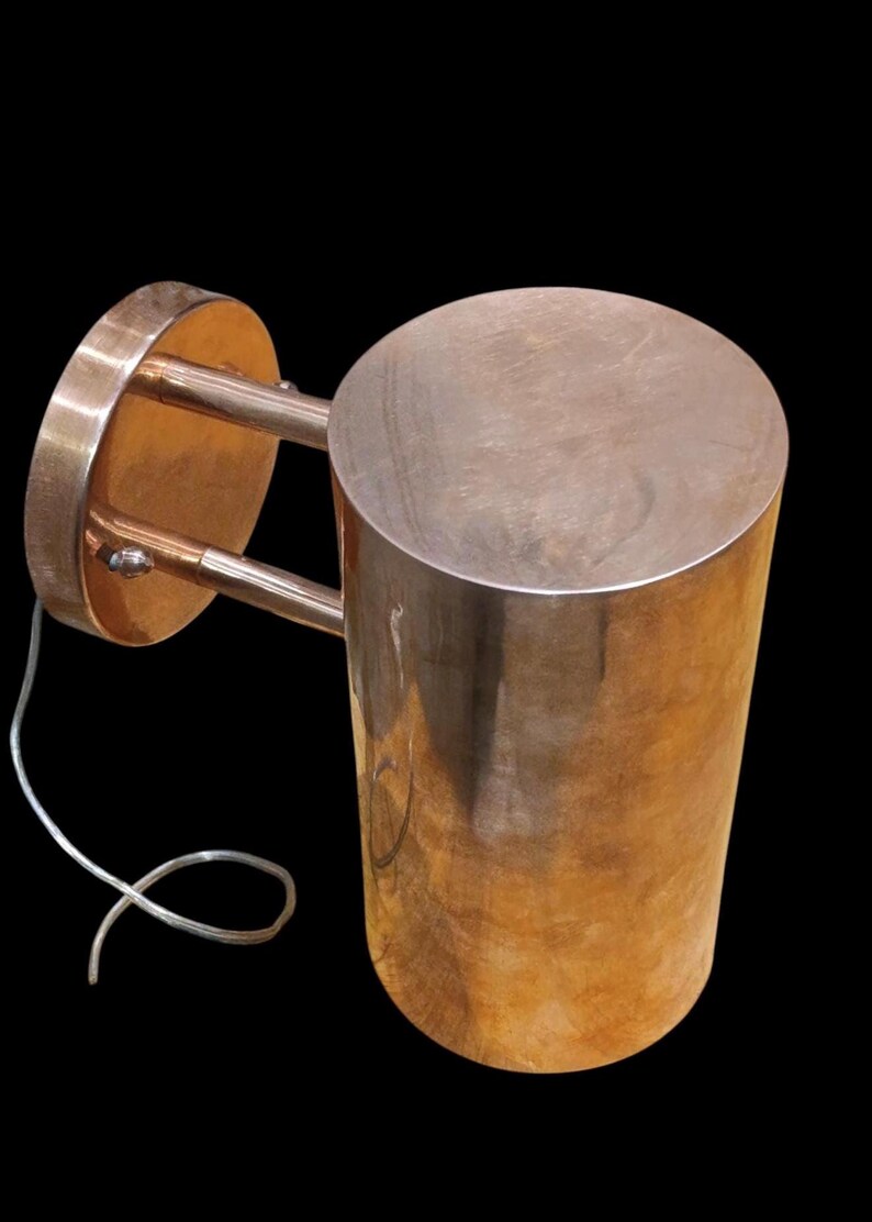May include: A copper-colored wall sconce with a cylindrical shade and a circular mounting plate. The sconce has two curved arms connecting the shade to the plate. A white electrical cord is visible.