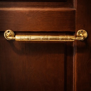 Puede incluir: Un tirador de puerta dorado con un diseño ornamentado y texturizado. El tirador está montado en una puerta de madera marrón oscuro. El tirador tiene forma cilíndrica con detalles decorativos y placas de montaje circulares en cada extremo.