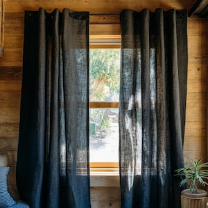 May include: Dark blue linen curtains hanging in front of a window. The curtains have a textured appearance and are partially blocking the sunlight. The room has wooden walls and a small wooden stool with a potted plant.