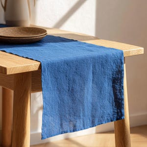 May include: A blue linen table runner draped over a wooden table. The runner has a textured appearance and is a deep shade of blue. A brown plate and a beige pitcher are visible on the table.