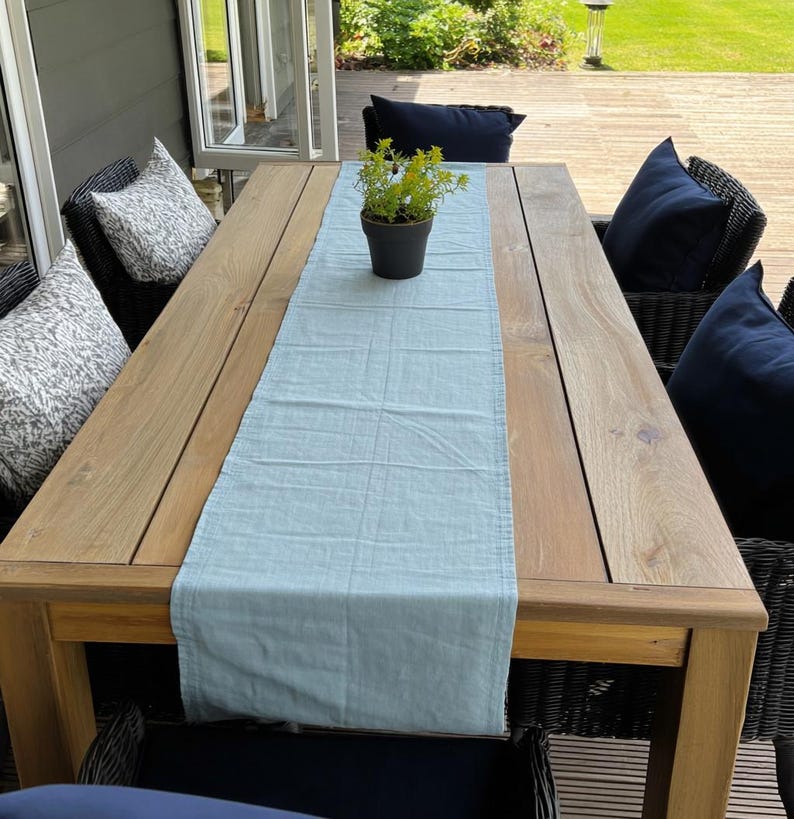 K&ouml;nnte beinhalten: Ein rechteckiger Esstisch aus hellem Holz im Freien mit einem hellblauen Tischl&auml;ufer. Der Tisch ist von schwarzen Korbst&uuml;hlen mit marineblauen Kissen und gemusterten Kissen umgeben. Auf dem Tisch steht eine kleine Topfpflanze.