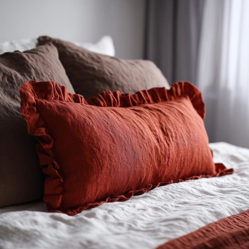 May include: A rectangular, rust-coloured pillow with frilled edges sits on a white bed. Two other pillows, one brown and one white, are behind it. The bedding has a textured appearance, possibly linen or a similar fabric.