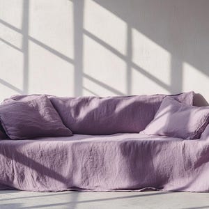 May include: A lavender-colored sofa with two matching throw pillows. The sofa is covered with a linen fabric, and the room has natural light from a window. The shadows from the window create a pattern on the wall.
