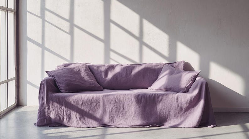 May include: A lavender-colored sofa with two matching throw pillows. The sofa is covered with a linen fabric, and the room has natural light from a window. The shadows from the window create a pattern on the wall.