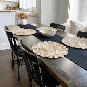 K&ouml;nnte beinhalten: Ein rustikaler Esstisch, gedeckt f&uuml;r eine Mahlzeit. Der dunkle Holztisch ist mit einem schwarzen Tischl&auml;ufer, gewebten Platzsets und einer Keramikschale geschm&uuml;ckt. Schwarze St&uuml;hle umgeben den Tisch.