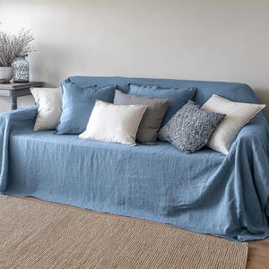 May include: A blue sofa covered with a matching linen throw and several decorative pillows. The pillows are in shades of white, gray, and blue, with various textures and patterns. A neutral-colored rug is in the foreground.