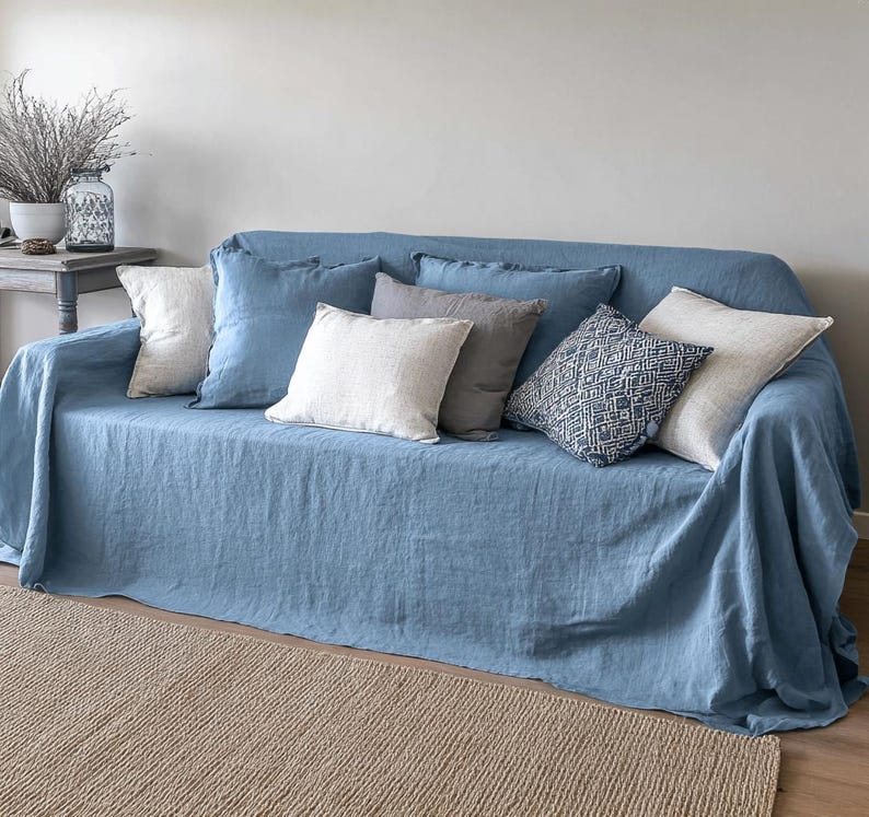 May include: A blue sofa covered with a matching linen throw and several decorative pillows. The pillows are in shades of white, gray, and blue, with various textures and patterns. A neutral-colored rug is in the foreground.