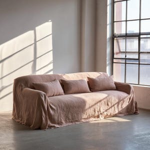 May include: A blush-colored sofa with three matching rectangular pillows. The sofa is covered in a draped fabric, and the room has a large window with sunlight streaming in. The overall aesthetic is minimalist and modern.
