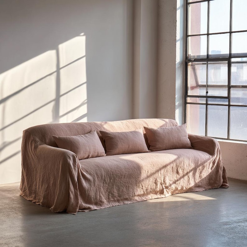 May include: A blush-colored sofa with three matching rectangular pillows. The sofa is covered in a draped fabric, and the room has a large window with sunlight streaming in. The overall aesthetic is minimalist and modern.