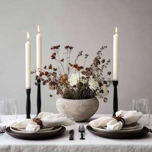May include: A dining table set with lit white candles in black holders, a stone vase with dried flowers, and place settings with white napkins and brown plates. The table is covered with a white linen tablecloth, creating a neutral and elegant aesthetic.