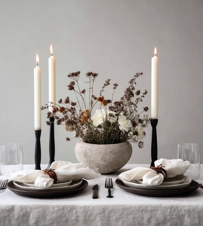May include: A dining table set with lit white candles in black holders, a stone vase with dried flowers, and place settings with white napkins and brown plates. The table is covered with a white linen tablecloth, creating a neutral and elegant aesthetic.