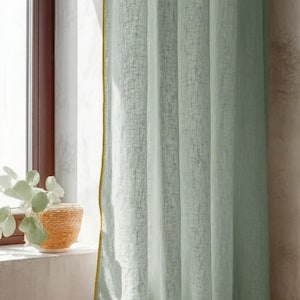 May include: Light green linen curtain with a yellow trim, hanging near a window. A woven basket with a plant sits on the windowsill. The curtain has a textured appearance and is partially lit by sunlight.