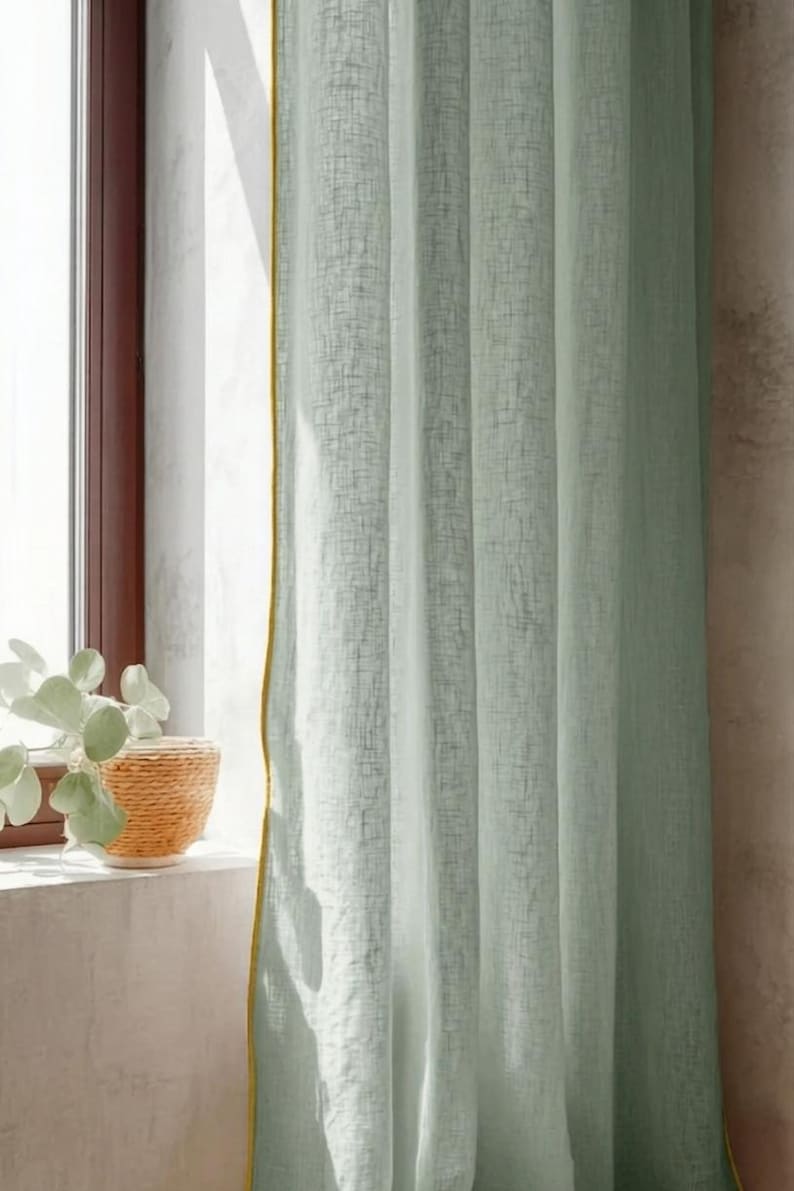 May include: Light green linen curtain with a yellow trim, hanging near a window. A woven basket with a plant sits on the windowsill. The curtain has a textured appearance and is partially lit by sunlight.