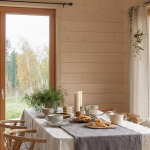 May include: A dining table set for a meal, featuring a white tablecloth with a gray runner. The table is set with plates, bowls, cups, and candles. Wooden chairs are positioned around the table, and a window offers a view of a natural landscape.