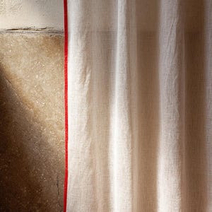 May include: Off-white linen curtain with a vibrant red trim. The curtain hangs in soft folds, showcasing the texture of the fabric. The red trim adds a bold contrast to the neutral color of the curtain.