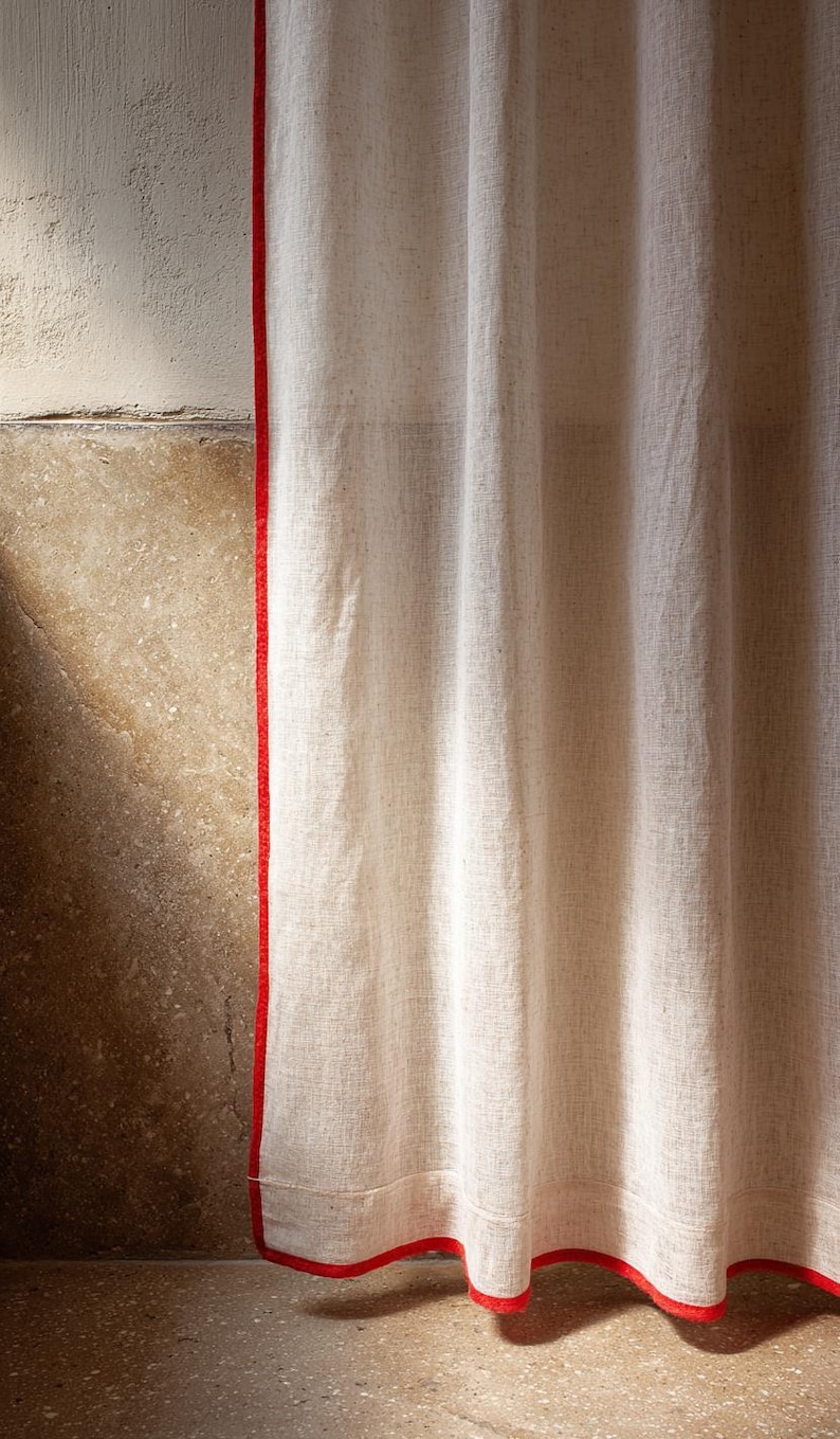 May include: Off-white linen curtain with a vibrant red trim. The curtain hangs in soft folds, showcasing the texture of the fabric. The red trim adds a bold contrast to the neutral color of the curtain.