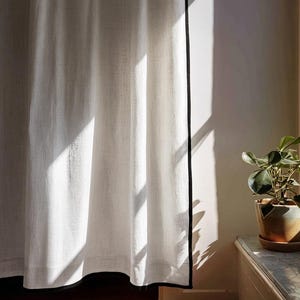 May include: White linen curtain with a black trim, illuminated by sunlight. The curtain hangs in soft folds, creating shadows. A potted plant sits on a nearby surface, adding a touch of greenery to the scene. The curtain is a home decor item.