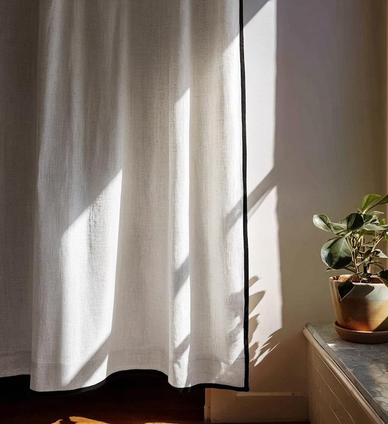 May include: White linen curtain with a black trim, illuminated by sunlight. The curtain hangs in soft folds, creating shadows. A potted plant sits on a nearby surface, adding a touch of greenery to the scene. The curtain is a home decor item.