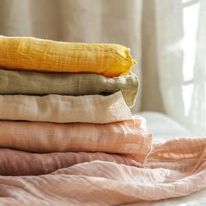 K&ouml;nnte beinhalten: Ein Stapel gefalteter Stoffe in verschiedenen Pastellfarben. Die Farben umfassen Gelb, Olivgr&uuml;n, Creme, Pfirsich und Altrosa. Der Stoff scheint leicht und strukturiert zu sein, m&ouml;glicherweise Leinen oder Gaze. Der Stapel liegt auf einer wei&szlig;en Oberfl&auml;che.