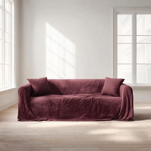 May include: A burgundy linen sofa with two matching throw pillows. The sofa is draped with a textured fabric, and the room has large windows and light-colored wood flooring. The overall aesthetic is minimalist and cozy.