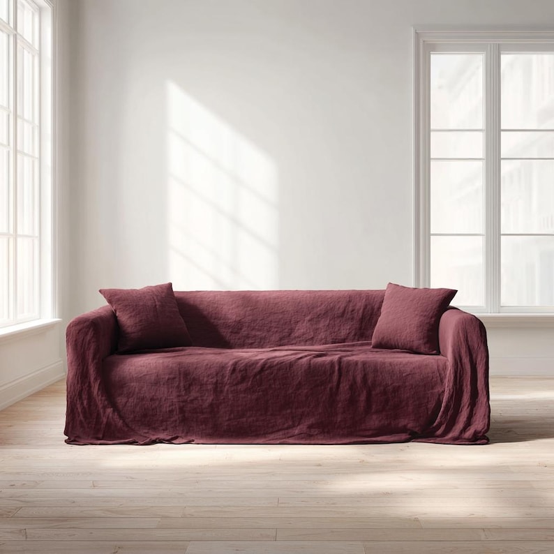 May include: A burgundy linen sofa with two matching throw pillows. The sofa is draped with a textured fabric, and the room has large windows and light-colored wood flooring. The overall aesthetic is minimalist and cozy.