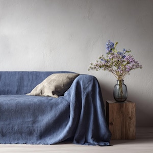 May include: A blue linen sofa is draped with a matching throw. A beige pillow rests on the sofa. A vase of purple and blue flowers sits on a wooden block, creating a cozy, inviting living space.