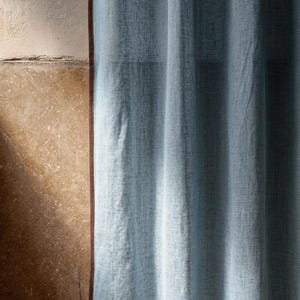 May include: A light blue linen curtain with a brown trim, hanging in front of a textured beige wall. The curtain is slightly gathered, and the fabric has a subtle woven pattern. The image is well-lit, with soft shadows.