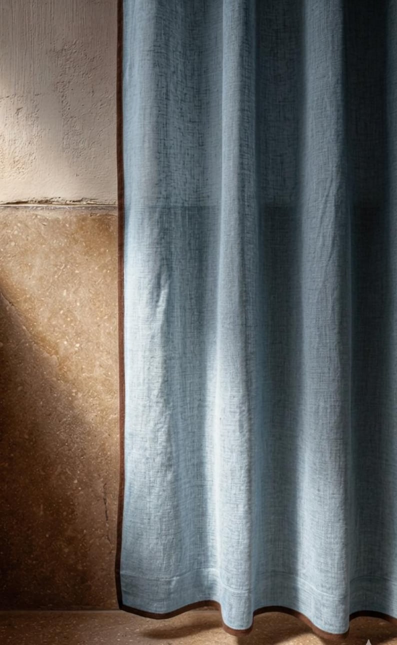 May include: A light blue linen curtain with a brown trim, hanging in front of a textured beige wall. The curtain is slightly gathered, and the fabric has a subtle woven pattern. The image is well-lit, with soft shadows.