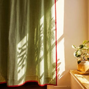 May include: A light green curtain with a red trim, casting shadows from a nearby plant. The curtain is made of a textured fabric and hangs in front of a white wall. A potted plant sits on a wooden surface.
